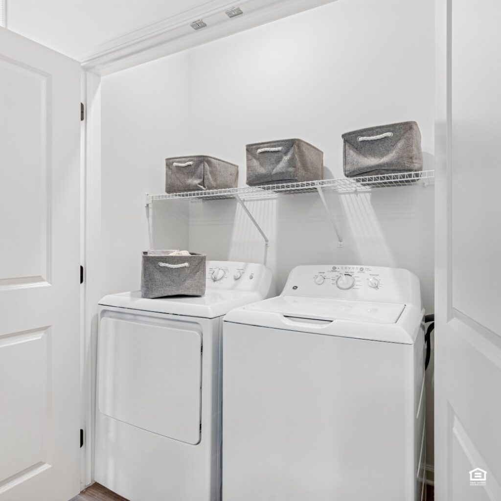 White laundry closet with top-load machines and gray storage bins