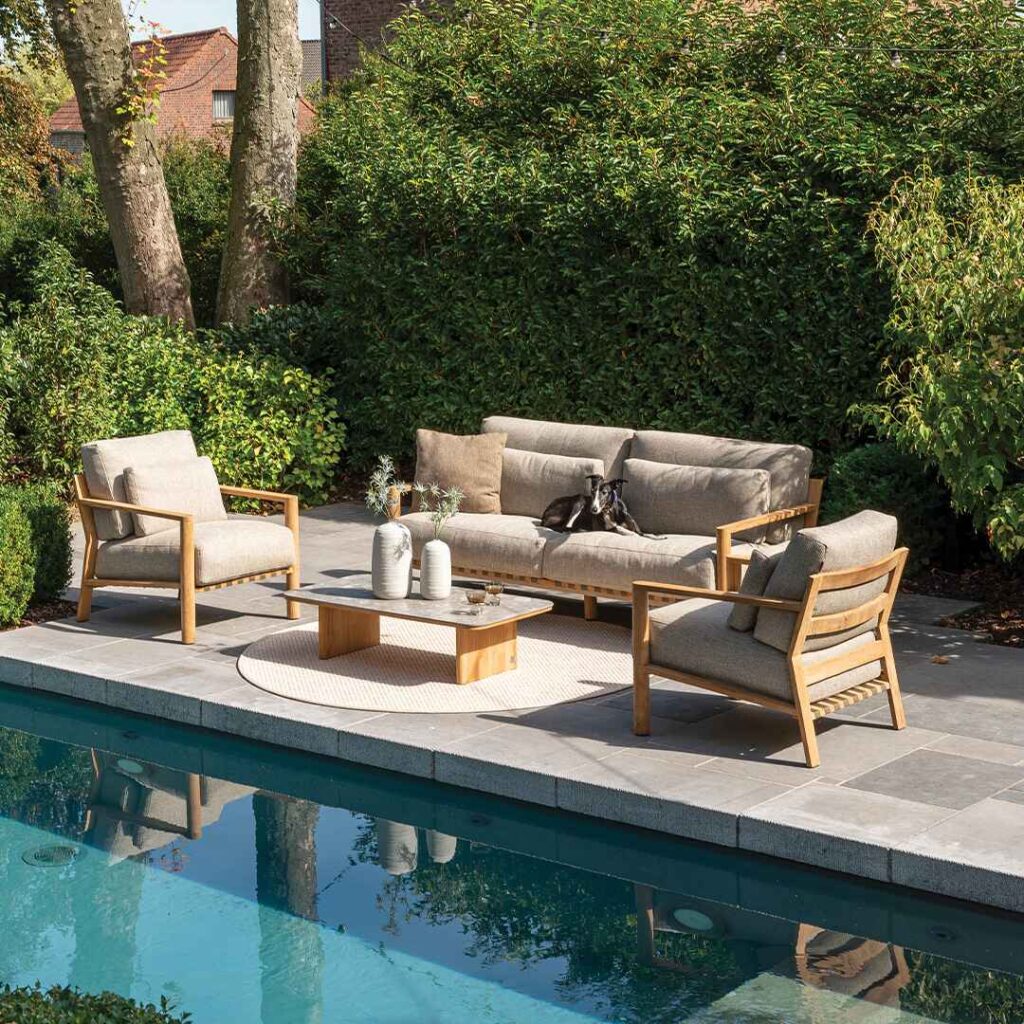 Modern wooden outdoor furniture set beside pool surrounded by lush greenery.