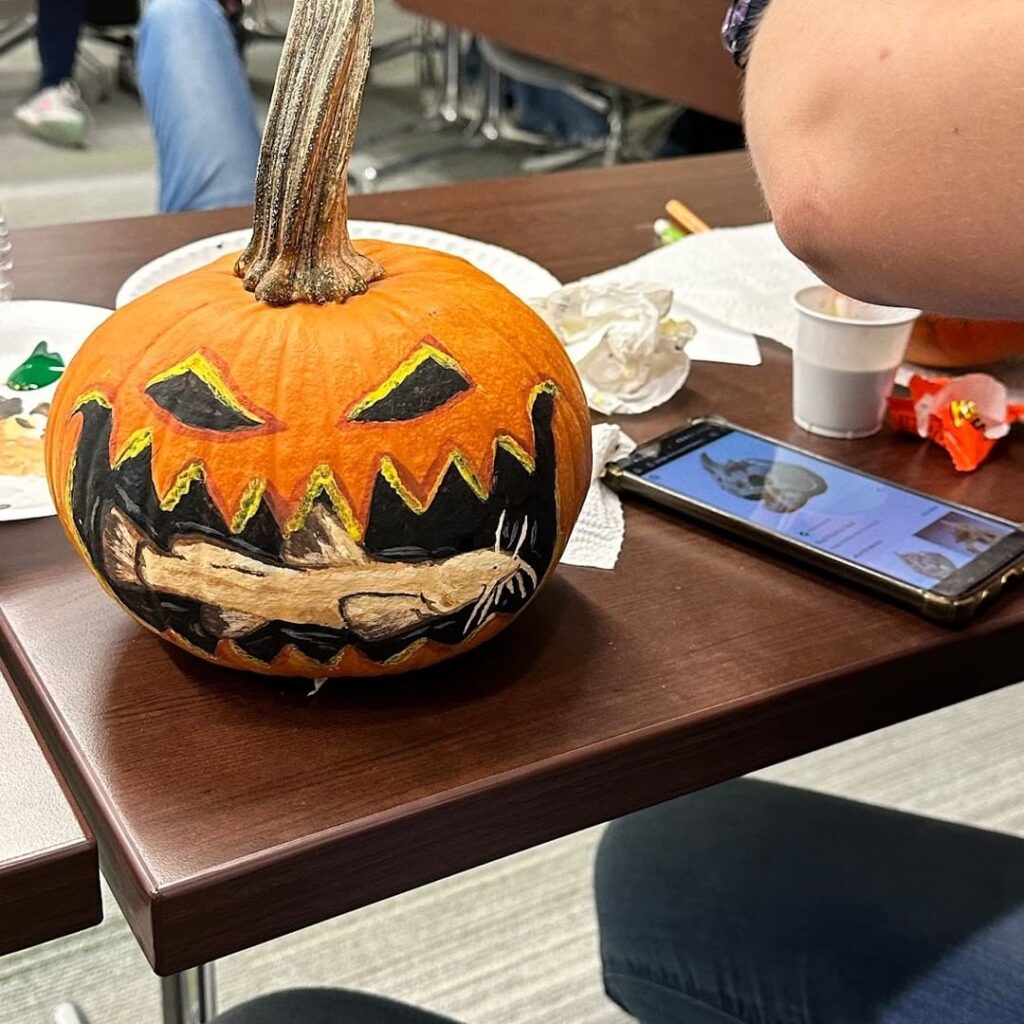 Painted pumpkin with grinning face holding bone in teeth.