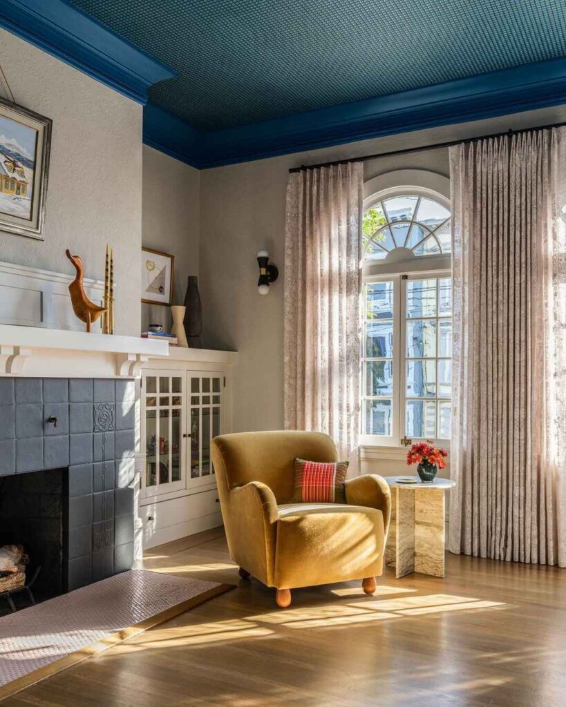 Sunlit living room corner with mustard chair fireplace and blue ceiling.