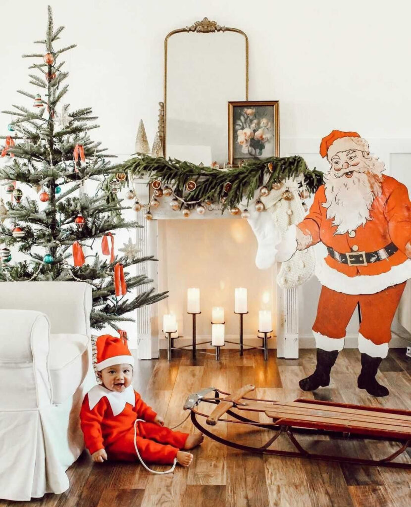 Baby in elf costume with Christmas tree, fireplace, and Santa cutout.