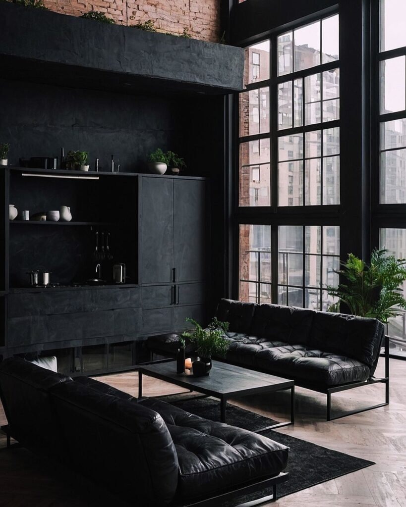 Dark loft living room with black furniture and large windows overlooking cityscape.