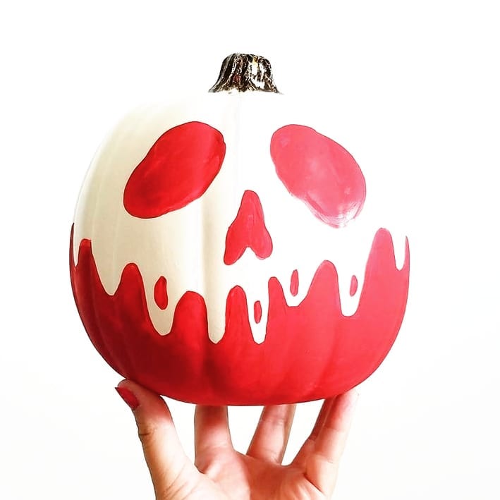Hand holding white pumpkin painted as red poison apple with skull face.