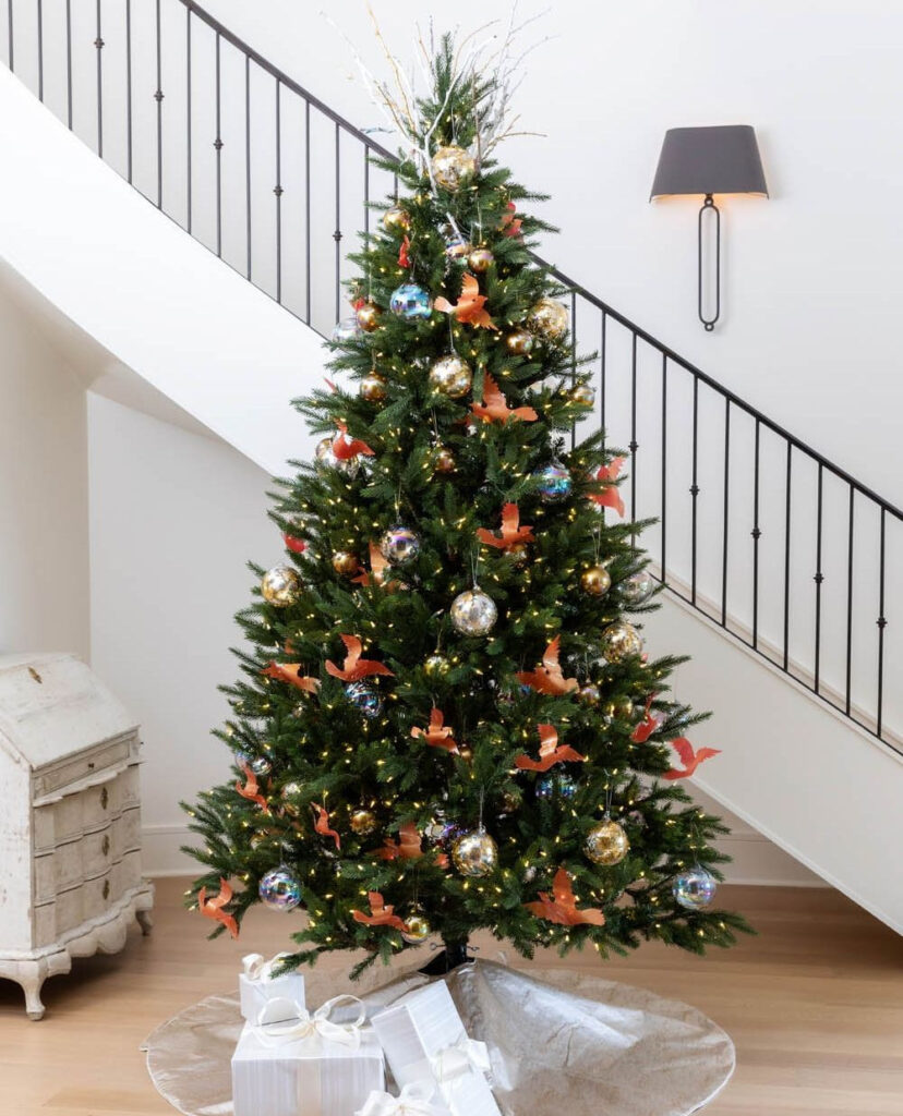 Christmas tree with orange bird ornaments and metallic baubles by staircase.