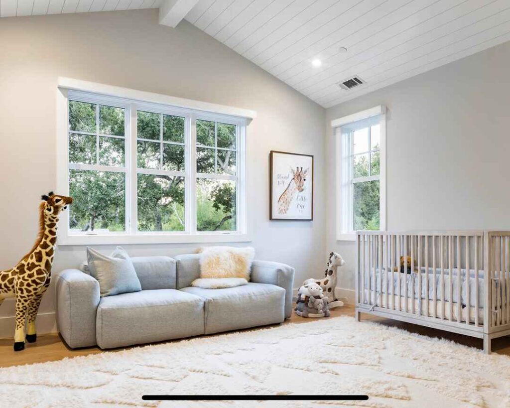 Modern nursery with gray sofa large giraffe plush and white crib near windows