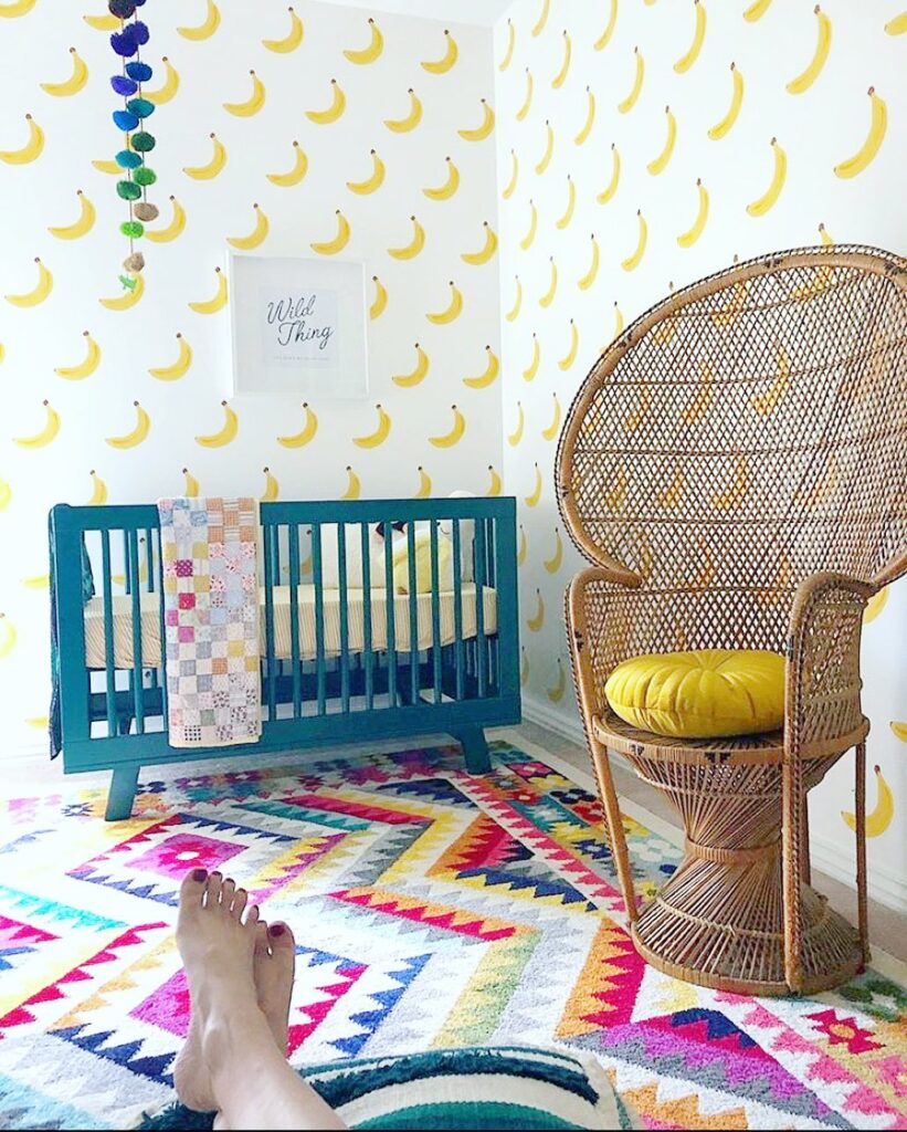 Nursery with banana print wallpaper teal crib and woven peacock chair on colorful rug